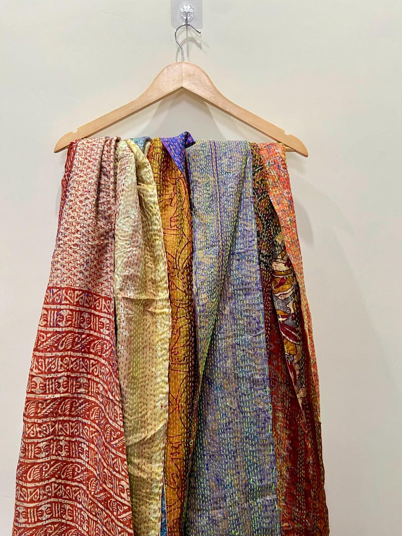 Op de afbeelding: Een verzameling kleurrijke, bedrukte sjaals die aan een houten kleerhanger hangen. De sjaals tonen een verscheidenheid aan kleuren, waaronder rood, geel, blauw en oranje, met ingewikkelde gestikte ontwerpen. De stoffen zijn gedrapeerd en tonen hun texturen en patronen.