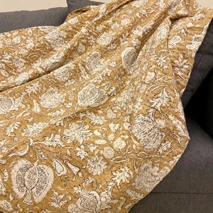 Peut inclure: Une couverture à motifs floraux de couleur moutarde drapée sur un canapé gris. La couverture présente un motif répété de grandes fleurs et de feuilles blanches. Le tissu semble être matelassé avec des coutures visibles.