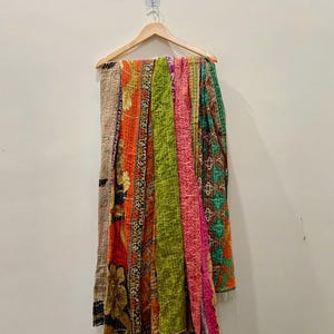 May include: A colorful, patchwork quilt hanging on a wooden hanger. The quilt features various patterned fabrics in shades of orange, green, pink, and brown. The quilt is made of stitched fabric.