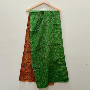 Op de afbeelding: Een groene en oranje sjaal hangt aan een houten hanger. De sjaal is gemaakt van een textuurstof. De groene kant heeft een gekreukeld uiterlijk, terwijl de oranje kant een patroon heeft.