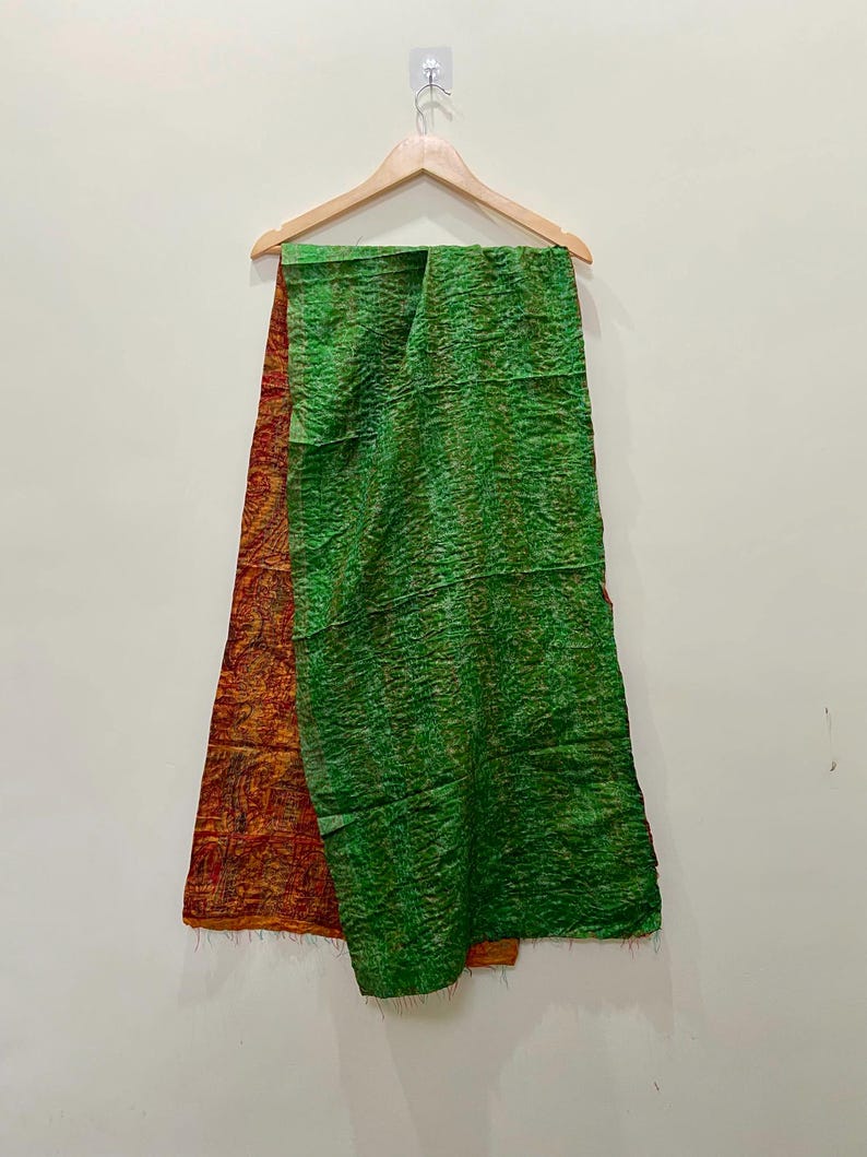 Op de afbeelding: Een groene en oranje sjaal hangt aan een houten hanger. De sjaal is gemaakt van een textuurstof. De groene kant heeft een gekreukeld uiterlijk, terwijl de oranje kant een patroon heeft.
