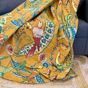 May include: Mustard-yellow textile featuring a detailed bird and floral pattern in red, white, blue, and green. The fabric is draped over a blue sofa and a natural fiber rug, highlighting the intricate design and colour palette.