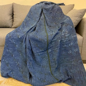 Könnte beinhalten: Eine blaue Decke mit einem gemusterten Design liegt über einem grauen Sofa. Die Decke hat eine genähte Textur mit einem grünen Streifen in der Mitte. Die Decke liegt auf einem beigen Teppich mit gewelltem Rand.