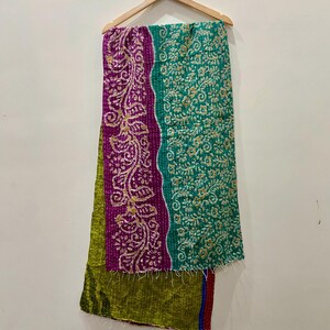 May include: A colorful textile hanging on a wooden hanger. The textile features a patchwork design with sections of green, purple, and teal. The purple section has a gold floral pattern. The textile is likely a scarf or a decorative piece.