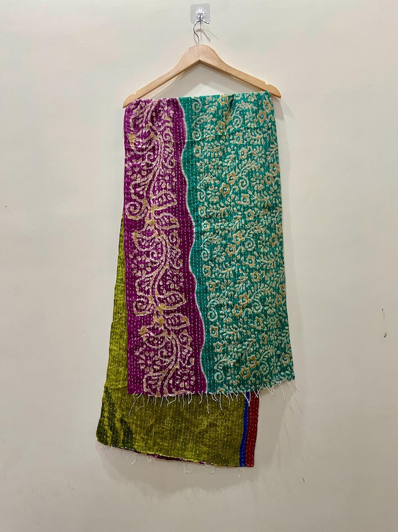May include: A colorful textile hanging on a wooden hanger. The textile features a patchwork design with sections of green, purple, and teal. The purple section has a gold floral pattern. The textile is likely a scarf or a decorative piece.