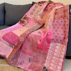 Peut inclure: Une couverture patchwork rose, allant du rose clair au rose foncé, drapée sur un canapé gris. La couverture présente divers carrés de tissu à motifs, notamment des motifs floraux et géométriques, cousus ensemble avec du fil visible.