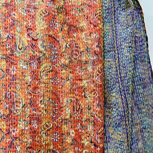 Op de afbeelding: Een levendig textiel met een paisley patroon in tinten oranje, geel en blauw. De stof is versierd met ingewikkelde stiksels en een complementaire blauwe en paarse stof is zichtbaar aan de rechterkant. Het textiel is waarschijnlijk een sjaal of een vergelijkbaar accessoire.