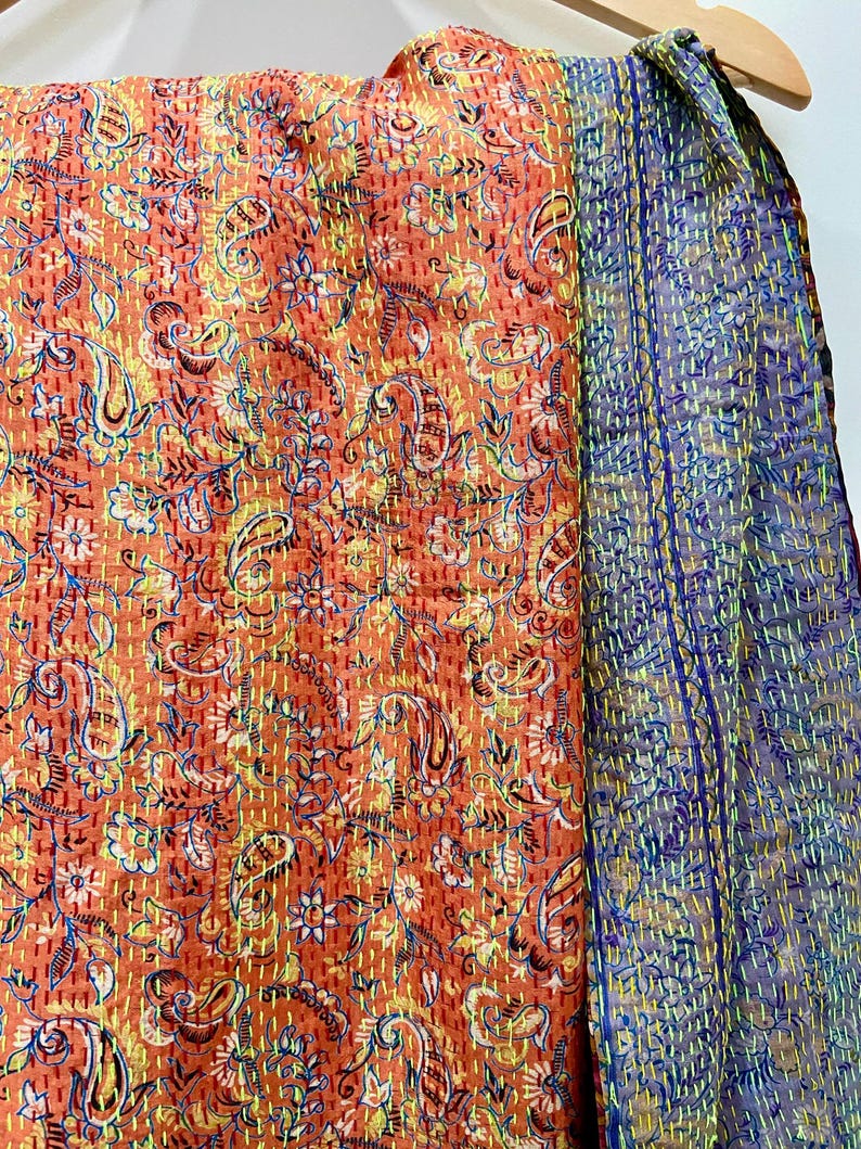 Op de afbeelding: Een levendig textiel met een paisley patroon in tinten oranje, geel en blauw. De stof is versierd met ingewikkelde stiksels en een complementaire blauwe en paarse stof is zichtbaar aan de rechterkant. Het textiel is waarschijnlijk een sjaal of een vergelijkbaar accessoire.