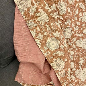 Op de afbeelding: Een gevouwen quilt met een bloemenpatroon in bruin- en cr&egrave;metinten. De achterkant is effen stoffig roze. De quilt is gestikt met witte draad, wat een textuur geeft. Gemaakt van katoen, ideaal voor bed of bank.