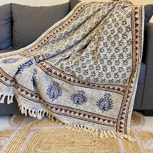 May include: A patterned throw blanket in beige and brown with a fringed edge is draped over a grey sofa. The blanket has a repeating floral design in black and blue, with a brown border.