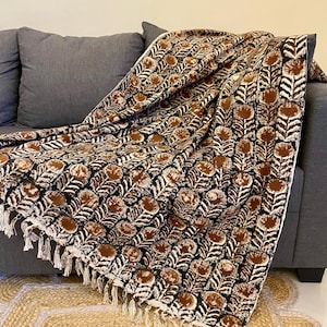 May include: A patterned throw blanket in shades of brown, black, and white, draped over a grey sofa. The blanket has a repeating floral design and a fringed edge. The blanket is made of a soft material and is suitable for home decor.