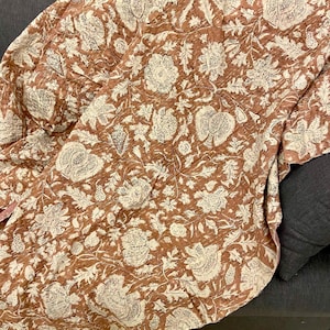 Op de afbeelding: Een terracotta kleurige quilt met een cr&egrave;mekleurig bloemenpatroon. De quilt is over een donkergrijze bank gedrapeerd. Het bloemmotief bevat grote bloemen, bladeren en andere botanische elementen. De quilt lijkt gemaakt van een zachte, gewatteerde stof.
