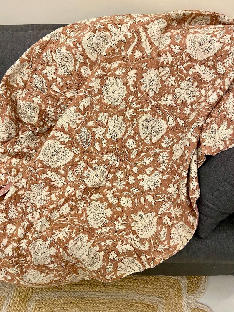Op de afbeelding: Een terracotta kleurige quilt met een cr&egrave;mekleurig bloemenpatroon. De quilt is over een donkergrijze bank gedrapeerd. Het bloemmotief bevat grote bloemen, bladeren en andere botanische elementen. De quilt lijkt gemaakt van een zachte, gewatteerde stof.