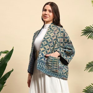 May include: A teal and beige patterned jacket with a geometric design. The jacket has long sleeves and two front pockets. The model is wearing a white top and a white skirt. A beige belt is worn at the waist.