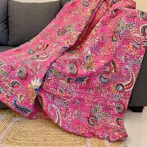 Peut inclure: Une couverture rose vif avec un motif floral détaillé dans les tons de bleu, jaune et rouge. La couverture est drapée sur un canapé gris, mettant en valeur son design et sa texture complexes. Le motif de la couverture comprend des éléments cachemire et floraux.