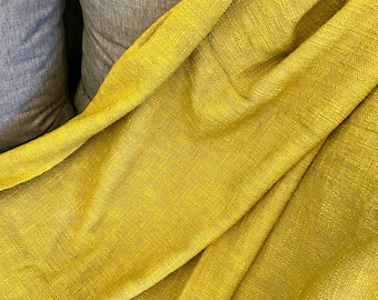 Puede incluir: Primer plano de una manta amarilla dorada texturizada sobre una almohada gris. La manta tiene un aspecto tejido y está doblada en curvas suaves y fluidas. El color es un tono vibrante y cálido.