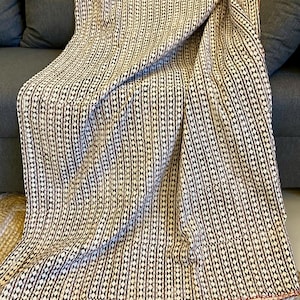 May include: A cream-colored throw blanket with a repeating pattern of vertical lines and diamond shapes in dark brown. The blanket is draped over a gray sofa, with a red stitched edge.