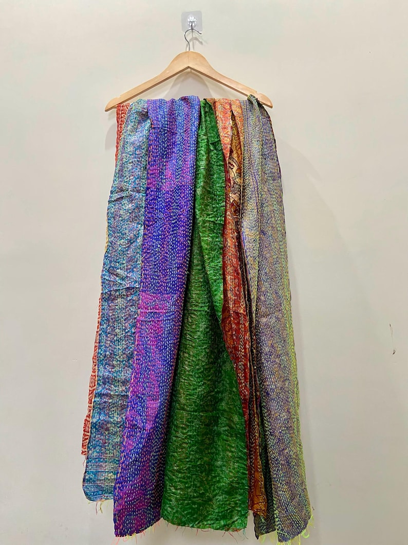 Op de afbeelding: Een verzameling kleurrijke, bedrukte sjaals hangt aan een houten kleerhanger. De sjaals tonen een verscheidenheid aan kleuren, waaronder blauw, paars, groen, oranje en geel. De stof lijkt getextureerd, met zichtbare stiksels.