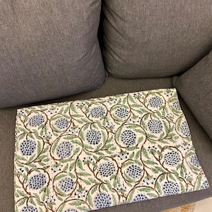 May include: A folded white fabric with a repeating pattern of blue floral clusters and green leaves connected by brown stems. The fabric is resting on a gray couch.