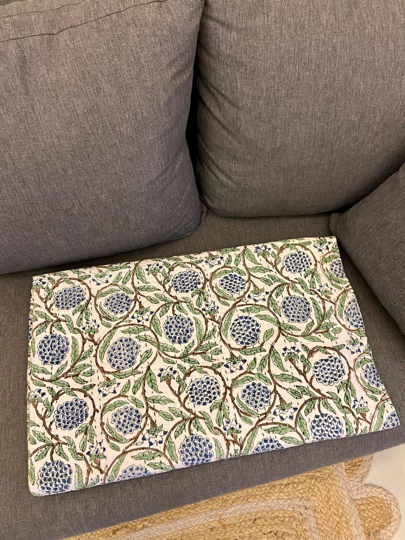 May include: A folded white fabric with a repeating pattern of blue floral clusters and green leaves connected by brown stems. The fabric is resting on a gray couch.