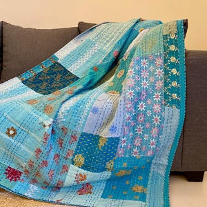 May include: A patchwork quilt in shades of blue, teal, and white, draped over a gray sofa. The quilt features floral and geometric patterns, with intricate stitching and a decorative border. The quilt is made of soft fabric.
