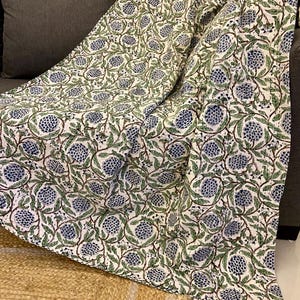 May include: A white blanket with a repeating pattern of blue floral designs and green leaves. The blanket is draped over a dark gray sofa. The blanket's edges are finished with a black border. The blanket is made of a soft, lightweight material.