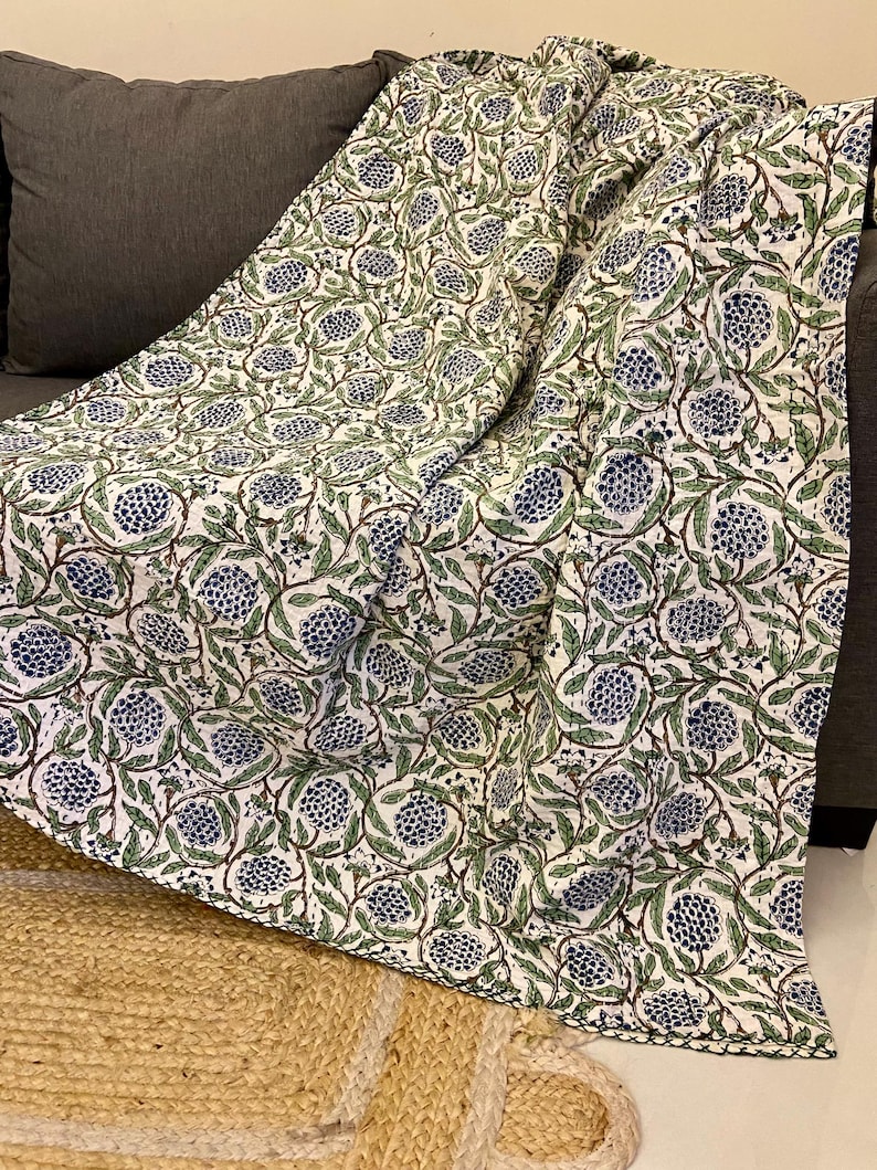 May include: A white blanket with a repeating pattern of blue floral designs and green leaves. The blanket is draped over a dark gray sofa. The blanket's edges are finished with a black border. The blanket is made of a soft, lightweight material.