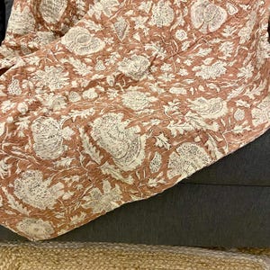Op de afbeelding: Een terracotta kleurige quilt met een bloemenpatroon in wit en cr&egrave;me. De quilt is over een donkergrijze ondergrond gevouwen, met een natuurvezel vloerkleed zichtbaar op de voorgrond. Het ontwerp van de quilt toont grote, gestileerde bloemen en bladeren.