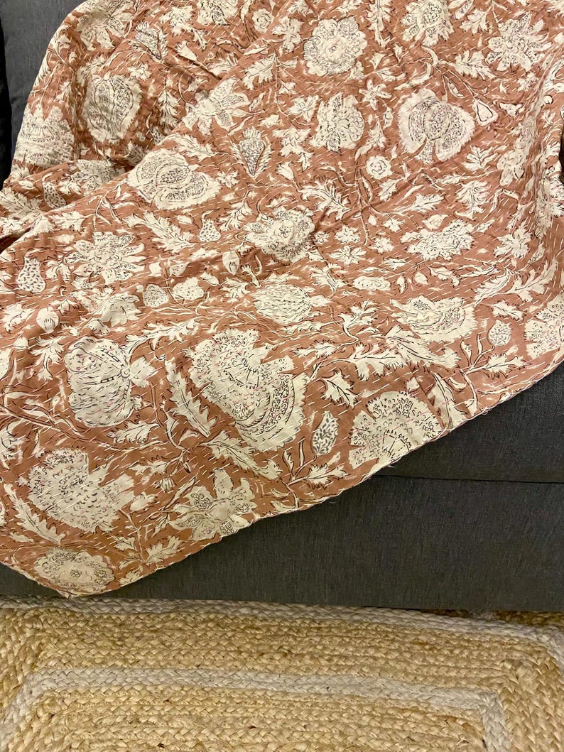 Op de afbeelding: Een terracotta kleurige quilt met een bloemenpatroon in wit en cr&egrave;me. De quilt is over een donkergrijze ondergrond gevouwen, met een natuurvezel vloerkleed zichtbaar op de voorgrond. Het ontwerp van de quilt toont grote, gestileerde bloemen en bladeren.