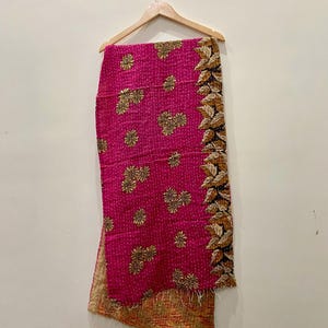 May include: A vibrant pink textile with gold floral embroidery and a decorative leaf border. The fabric is hanging from a wooden hanger, showcasing its intricate design and texture. The textile's reverse side has a warm, earthy tone.