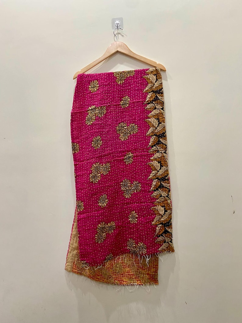 May include: A vibrant pink textile with gold floral embroidery and a decorative leaf border. The fabric is hanging from a wooden hanger, showcasing its intricate design and texture. The textile's reverse side has a warm, earthy tone.
