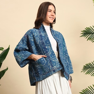 May include: A blue patterned jacket with a white and blue design. The jacket has a shawl collar, pockets, and a belt. The jacket is worn over a white turtleneck and skirt. The jacket is a medium weight and appears to be made of cotton.