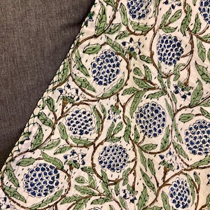 May include: Close-up of a white fabric with a repeating floral pattern. The design features clusters of blue flowers, green leaves, and brown vines. The fabric is stitched with white thread, creating a quilted effect. The background is a solid gray.