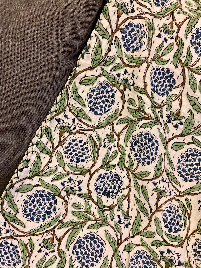 May include: Close-up of a white fabric with a repeating floral pattern. The design features clusters of blue flowers, green leaves, and brown vines. The fabric is stitched with white thread, creating a quilted effect. The background is a solid gray.