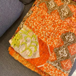 以下が含まれることがあります： オレンジ、茶色、クリーム色の鮮やかなパッチワークの織物。 花柄や幾何学模様など、複雑な模様が施され、ステッチが見えます。 織物は灰色の表面に置かれており、毛布またはスローを連想させます。