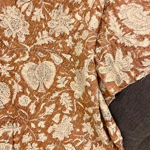Op de afbeelding: Een bruin en cr&egrave;mekleurig textiel met bloemenpatroon. De stof heeft een herhalend ontwerp van grote bloemen en bladeren, met witte stiksels. Het textiel is opgevouwen en onthult het patroon en de textuur.