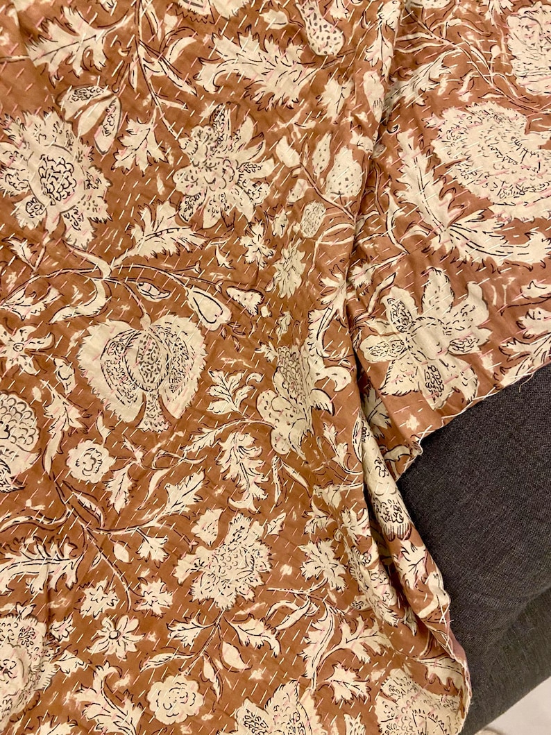 Op de afbeelding: Een bruin en cr&egrave;mekleurig textiel met bloemenpatroon. De stof heeft een herhalend ontwerp van grote bloemen en bladeren, met witte stiksels. Het textiel is opgevouwen en onthult het patroon en de textuur.