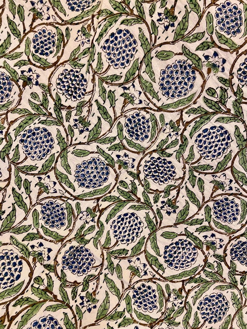 May include: Close-up of a fabric with a repeating floral pattern. The design features clusters of blue flowers, green leaves, and brown stems against an off-white background. The pattern is likely used for textiles or home decor.