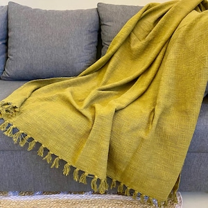 Puede incluir: Una manta tejida de color amarillo mostaza con flecos, colocada sobre un sofá gris. La manta tiene un aspecto texturizado y está hecha de un material suave. La manta es un textil decorativo para el hogar.