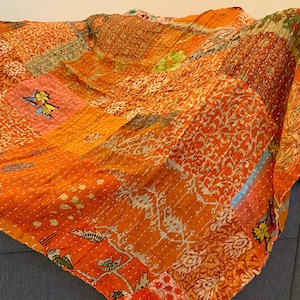 Pode incluir: Uma colcha de retalhos laranja com vários padrões e texturas. A colcha apresenta uma mistura de desenhos florais e geométricos em tons de laranja, dourado e branco. A colcha está sobre uma superfície cinza.