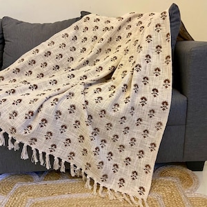 以下が含まれることがあります： 茶色の花柄が繰り返されるベージュ色のスローケットが、グレーのソファに掛けられています。ブランケットにはフリンジの縁取りがあります。幾何学模様の天然繊維ラグが手前にあります。