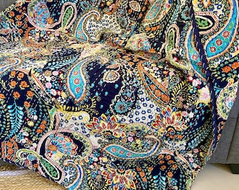 May include: A navy blue paisley patterned quilt with a vibrant mix of colors including teal, orange, yellow, and pink. The quilt is draped over a gray sofa and a natural fiber rug. The intricate design features floral and teardrop shapes.