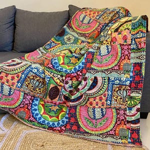 May include: A vibrant patchwork quilt with a mix of pink, blue, green, and yellow circular patterns, draped over a grey sofa. The quilt features floral and geometric designs and rests on a natural fibre rug.