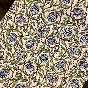 May include: Cream-colored fabric with a repeating pattern of blue floral clusters, green leaves, and brown vines. The design is printed on the fabric, creating a visually appealing and detailed textile. The overall aesthetic is reminiscent of a botanical print.