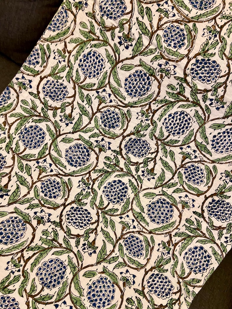 May include: Cream-colored fabric with a repeating pattern of blue floral clusters, green leaves, and brown vines. The design is printed on the fabric, creating a visually appealing and detailed textile. The overall aesthetic is reminiscent of a botanical print.
