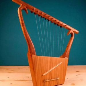 May include: A wooden lyre with a trapezoidal body and curved arms. The instrument has multiple strings stretched across the body and a horizontal bar. The lyre is made of light-coloured wood and sits on a wooden surface against a teal background.