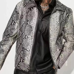 Puede incluir: Chaqueta con estampado de piel de serpiente en tonos grises y blancos. La chaqueta tiene cierre de cremallera y cuello. Se lleva sobre una camisa negra abotonada. El estilo general es atrevido y moderno.