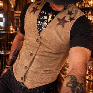 May include: A tan suede vest featuring dark brown star-shaped leather appliqués on the chest, a button-front closure with four brown buttons, and two front welt pockets. The vest is worn over a black t-shirt with white text that reads FLORIDA.