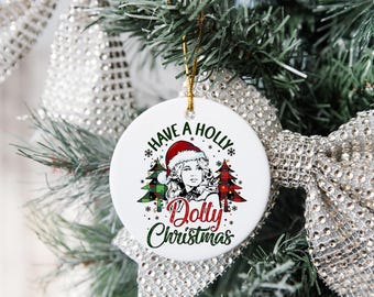 May include: A white ceramic Christmas ornament with a gold string. The ornament features a portrait of a woman wearing a Santa hat, with the text "Have a Holly Dolly Christmas" in green and red. Two Christmas trees flank the portrait.