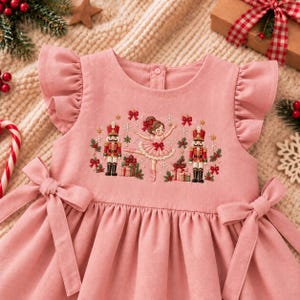 May include: A dusty pink sleeveless dress featuring ruffled shoulders and side waist ties. The bodice displays an embroidered scene of a ballerina dancing between two nutcrackers, surrounded by gift boxes, holly, and snowflakes in red and green.